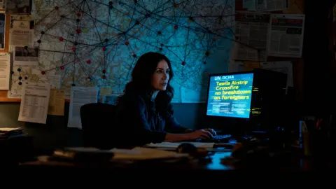 Still from No Splash: a woman working late at a desk in front of a wall of maps and notes