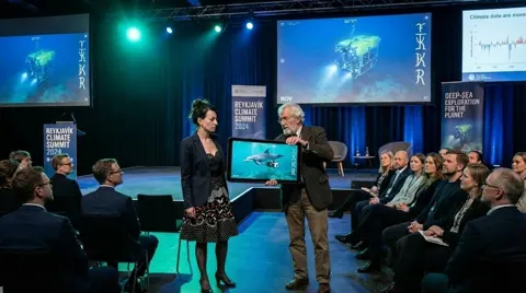 Still from The Sea Candle: Diddy Wheldon on stage at the Reykjavik summit beside a scientist with a tablet