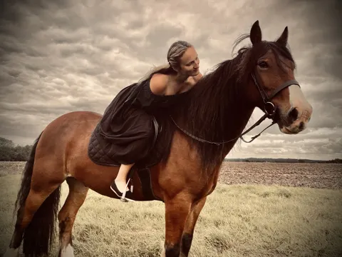 Still from Trapped in Their Dreams: a woman in a dark dress sitting on a bay horse under a cloudy sky
